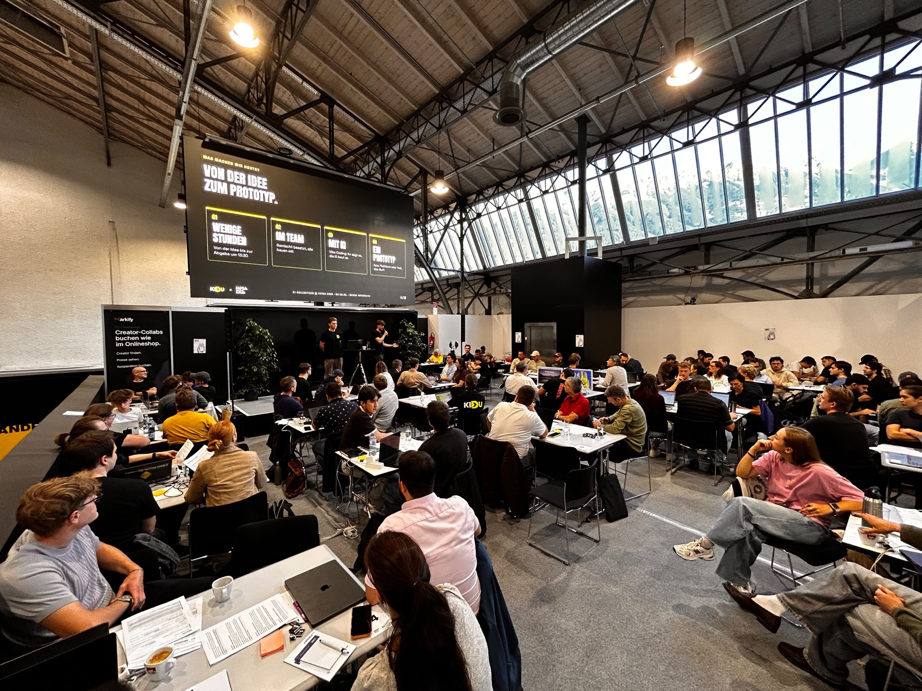 Blick in die volle Messehalle: rund achtzig Teilnehmende sitzen an Arbeitstischen, vorne die Bühne mit zwei Personen, darüber die große Leinwand mit dem Ablauf des Tages