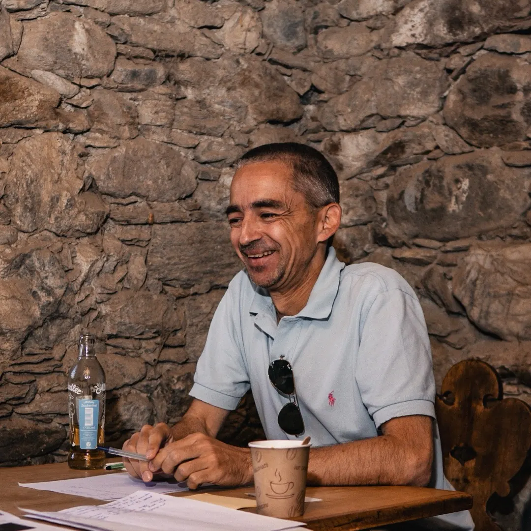 Ein Mann Mitte fünfzig im Poloshirt sitzt lachend an einem Holztisch vor einer Steinwand, den Stift in der Hand, vor ihm Notizzettel und ein Pappbecher.